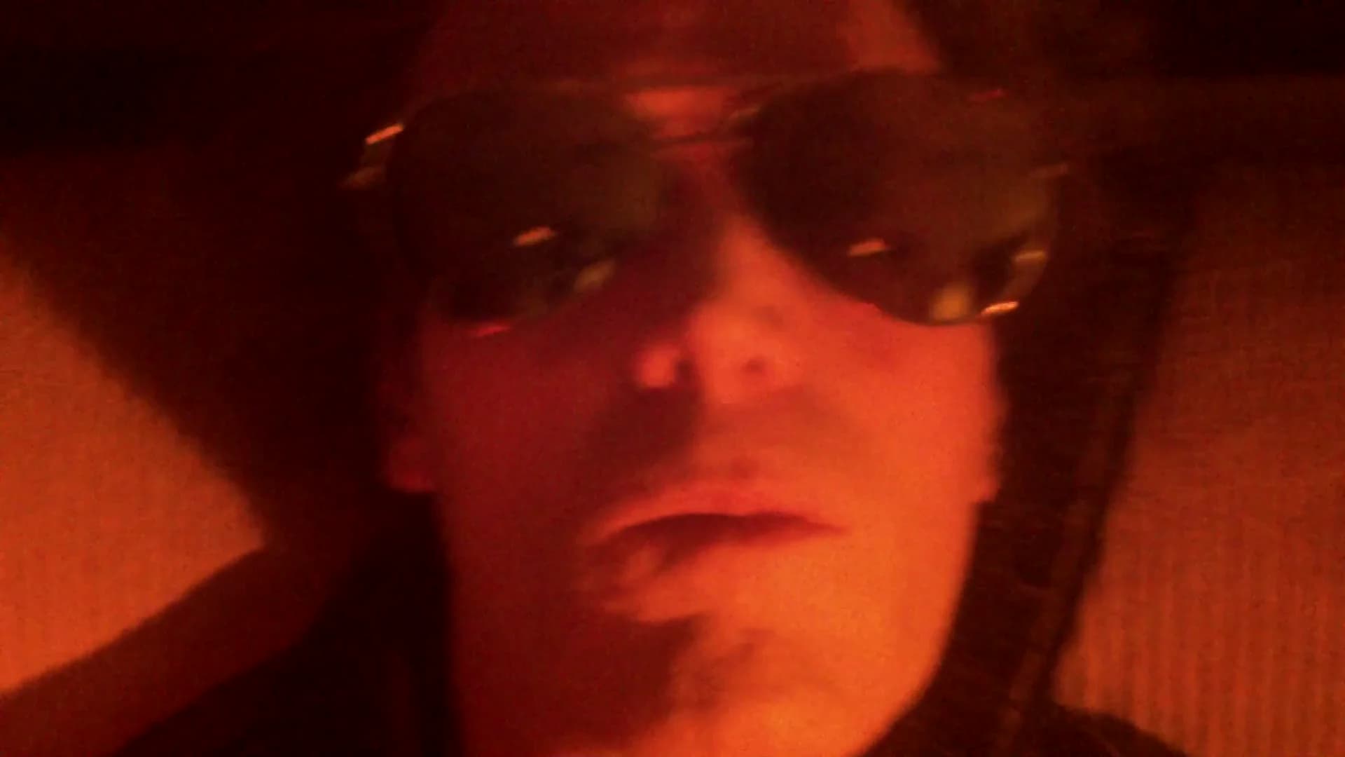 A pilot in dark glasses in the red light of a spacecraft cockpit.